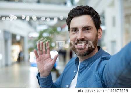 Cheerful man with a beard taking a selfie while waving in the bustling environment of a busy shopping center. Cheerful man with a beard taking a selfie while waving in the bustling environment of a busy shopping center. 112675829