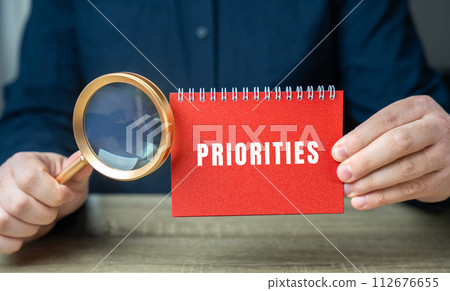 Note with the word Priorities in male hands. Concept showing importance, primacy. Magnifying glass Note with the word Priorities in male hands. Concept showing importance, primacy. Magnifying glass 112676655