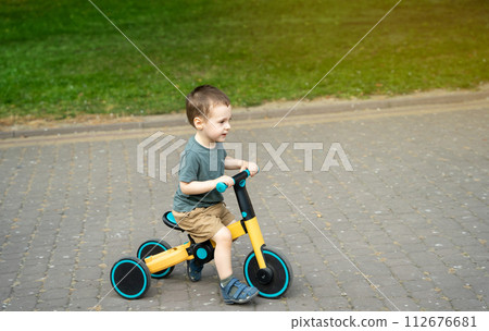A smiling toddler boy of two or three years old rides a bicycle or balance bike in a city park on a sunny summer day. Toddlerhood and childhood concept. Selective focus A smiling toddler boy of two or three years old rides a bicycle or balance bike in a city park on a sunny summer day. Toddlerhood and childhood concept. Selective focus 112676681