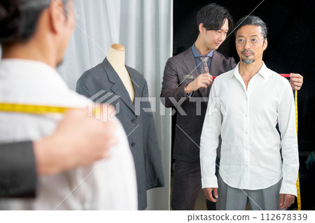 An elderly man looking in the mirror and a young clerk taking measurements in a fitting room 112678339