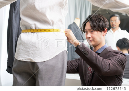 Male tailor staff taking customer's measurements in front of the mirror 112678343