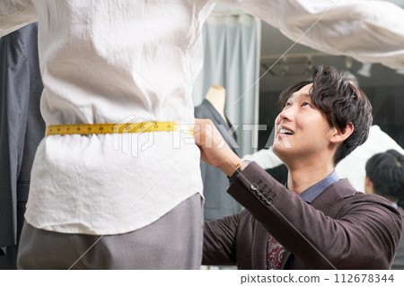 Male tailor staff taking customer's measurements in front of the mirror 112678344