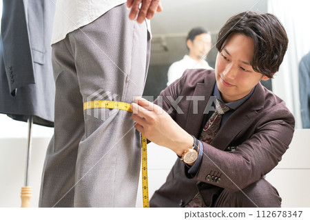 Male tailor staff taking customer's measurements in front of the mirror 112678347