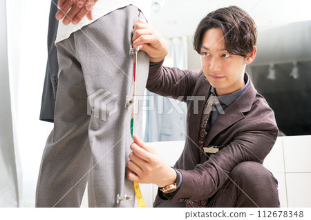 Male tailor staff taking customer's measurements in front of the mirror Male tailor staff taking customer's measurements in front of the mirror 112678348