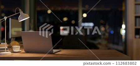 A laptop in a contemporary office room at night illuminated by dim lights from a table lamp. 112683802