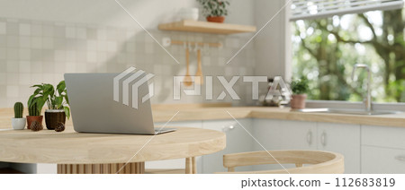 A back view image of a laptop computer on a wooden dining table in a minimalist neutral kitchen. 112683819