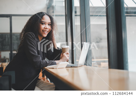 Confident young entrepreneur smiling while working at her laptop in a sunny coffee shop, illustrating successful remote work. 112684144