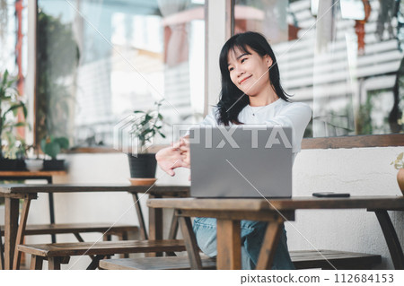Relaxed young woman stretching while working on a laptop in a casual outdoor cafe setting, enjoying a work-life balance. Relaxed young woman stretching while working on a laptop in a casual outdoor cafe setting, enjoying a work-life balance. 112684153