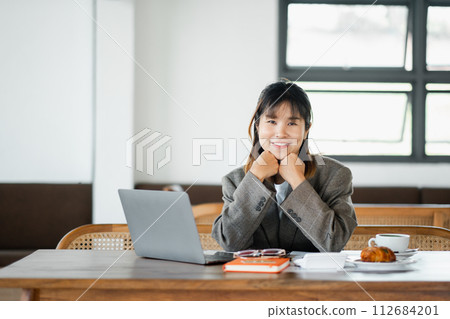 Content professional woman smiling at camera, with hands under chin, sitting at her workstation with laptop and breakfast on the table. 112684201