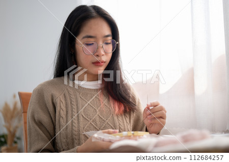 A young Asian woman is focusing on threading a pattern on an embroidery frame, hand sewing on cloth. 112684257