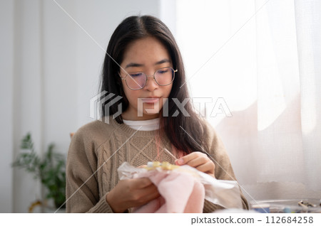 A young Asian woman is focusing on threading a pattern on an embroidery frame, hand sewing on cloth. 112684258