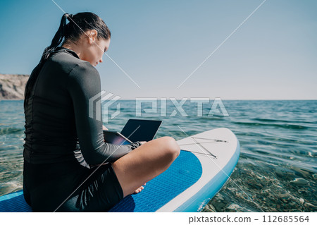 Woman sea laptop. Digital nomad, freelancer with laptop working on sup board at calm sea beach. Happy smiling girl relieves stress from work. Freelance, digital nomad, travel and holidays concept 112685564