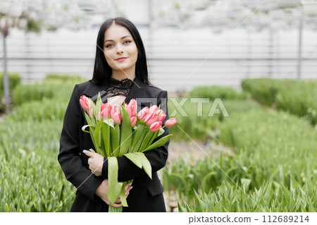 Young woman standing in large greenhouse and holding a blooming tulips in her hands Young woman standing in large greenhouse and holding a blooming tulips in her hands 112689214