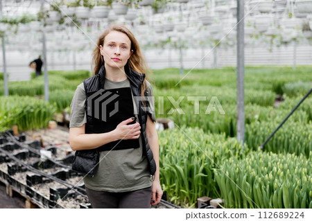 Young female worker with copybook standing in greenhouse with tulips 112689224