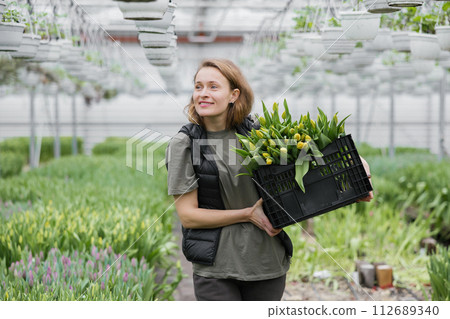 Young woman standing in greenhouse with box with tulips. Concept work in the greenhouse Young woman standing in greenhouse with box with tulips. Concept work in the greenhouse 112689340