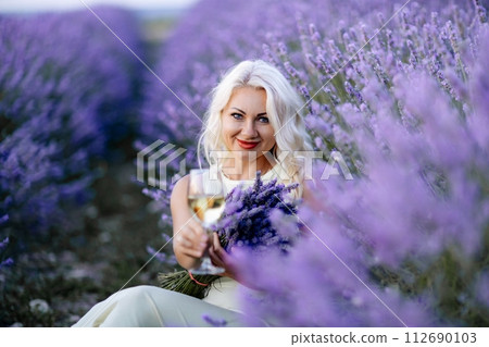 Blonde lavender field holds a glass of white wine in her hands. Happy woman in white dress enjoys lavender field picnic holding a large bouquet of lavender in her hands . Illustrating woman's picnic Blonde lavender field holds a glass of white wine in her hands. Happy woman in white dress enjoys lavender field picnic holding a large bouquet of lavender in her hands . Illustrating woman's picnic 112690103