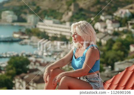 Woman sits on rooftop, enjoys town view and sea mountains. Peaceful rooftop relaxation. Below her, there is a town with several boats visible in the water. Rooftop vantage point. Woman sits on rooftop, enjoys town view and sea mountains. Peaceful rooftop relaxation. Below her, there is a town with several boats visible in the water. Rooftop vantage point. 112690106