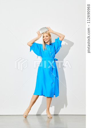 Cheerful woman in a blue shirtdress holds a silver purse over her head 112690968