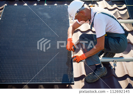Worker building photovoltaic solar panel system on rooftop of house. Man engineer in gloves installing solar module with help of hex key outdoors. Alternative and renewable energy generation concept. Worker building photovoltaic solar panel system on rooftop of house. Man engineer in gloves installing solar module with help of hex key outdoors. Alternative and renewable energy generation concept. 112692145