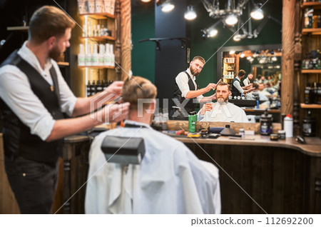 Regular client enjoying his drink while getting haircut. Professional hairdresser making stylish haircut, Luxury barbershop offering drink. Regular client enjoying his drink while getting haircut. Professional hairdresser making stylish haircut, Luxury barbershop offering drink. 112692200