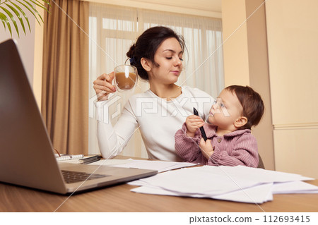 Cheerful businesswoman working at home with her little child girl and drinking coffee Cheerful businesswoman working at home with her little child girl and drinking coffee 112693415