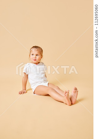 Joyful little baby, pretty girl in white romper sitting on floor with cute cheerful smile against beige studio background. Joyful little baby, pretty girl in white romper sitting on floor with cute cheerful smile against beige studio background. 112695060