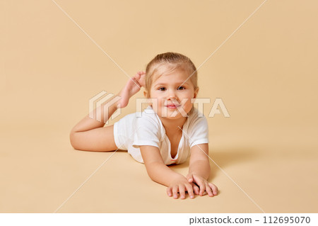 Cute little baby with curly resting on floor looking at camera with curious expression against pastel color background. Cute little baby with curly resting on floor looking at camera with curious expression against pastel color background. 112695070
