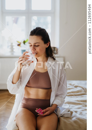 Woman taking pills with water, suffering from menstrual pain, having cramps. Close up of woman holding abdomen, endometriosis, and conditions causing pain in tummy. Woman taking pills with water, suffering from menstrual pain, having cramps. Close up of woman holding abdomen, endometriosis, and conditions causing pain in tummy. 112695494