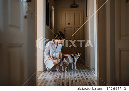 Woman in pajamas playing with her cute dog, drinking cup of morning coffee. Weekend morning relaxation at home. Hygge lifestyle. 112695540