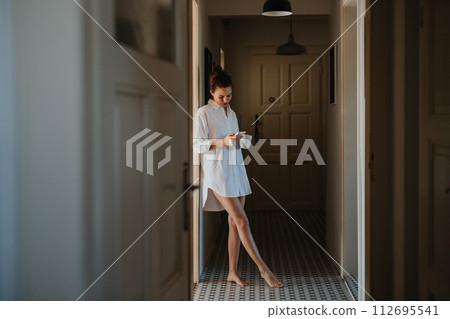 Young woman in the morning, carrying cup of coffee and returning back to bed. Side view of beautiful single woman at home alone over the weekend. Young woman in the morning, carrying cup of coffee and returning back to bed. Side view of beautiful single woman at home alone over the weekend. 112695541
