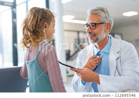 Little girl listening to the doctor's heart with stethoscope. Role reversal. Friendly relationship between the doctor and child patient. Little girl listening to the doctor's heart with stethoscope. Role reversal. Friendly relationship between the doctor and child patient. 112695546