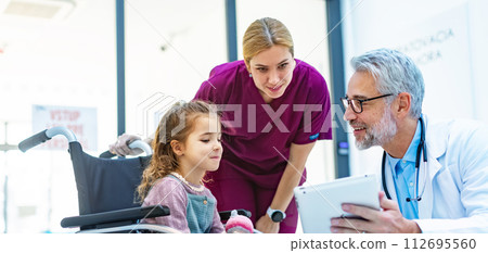 Friendly pediatrician showing someting on tablet to little patient in wheelchair. Cute preschool girl trusting her doctor and nurse in hospital. 112695560