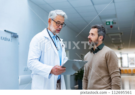 Doctor talking with patient in hospital corridor. Handsome doctor with gray hair holding clipboard with test result, consulting with man, standing in modern clinic. 112695561