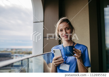 Nurse, doctor in uniform taking break, standing on hospital balcony, drinking coffee, scrolling on smartphone. Work-life balance of healthcare worker. 112695582