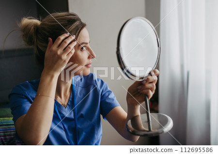 Female nurse or doctor getting ready for work in the morning, brushing hair, making hairstyle in bedroom. Work-life balance for healthcare worker. 112695586