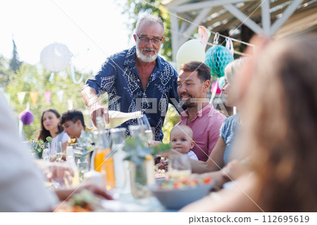 Grandfather opening bottle of champagne, pouring wine into glasses. Senior man making celebratory toast at outdoor summer garden party. Grandfather opening bottle of champagne, pouring wine into glasses. Senior man making celebratory toast at outdoor summer garden party. 112695619