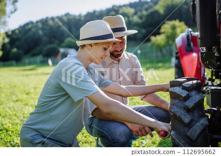 Farmer teaching son, how to fix wheel on tractor. Harvesting crops, collecting vegetables on family farm. Concept of farming, agricultural business. 112695662