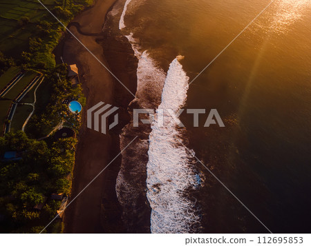 Aerial view of black sand beach with sunrise or sunset tones and ocean with waves in Bali Aerial view of black sand beach with sunrise or sunset tones and ocean with waves in Bali 112695853