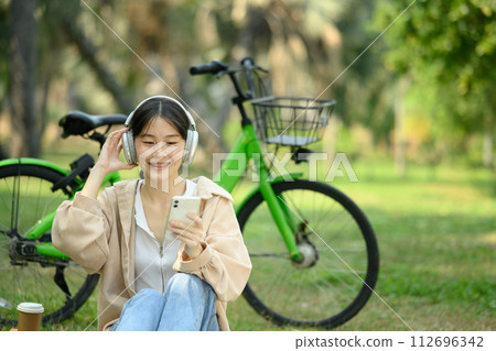 Smiling joyful young woman listening music over smartphone sitting on grass near her bicycle 112696342