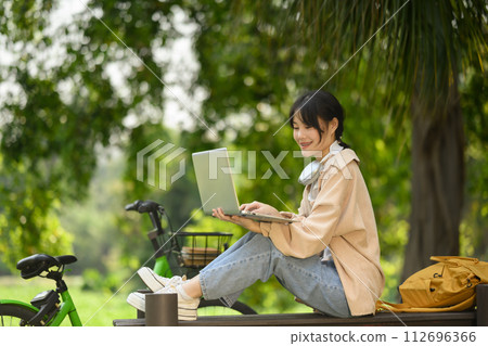 Smiling Asian female student using laptop, sitting on wooden bench in the park 112696366