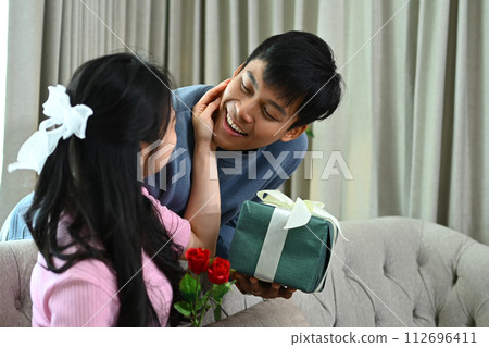 Smiling man surprising his girlfriend with a gift box on couch at home. Love, family and celebration concept 112696411