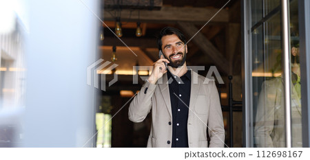 Portrait of mature businessman in suit jacket standing indoors in modern office, making phone call. Portrait of mature businessman in suit jacket standing indoors in modern office, making phone call. 112698167