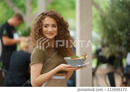 Portrait of a beautiful waitress holding plate with food, bowl with salad. Server standing on restaurant terrace in an apron. Portrait of a beautiful waitress holding plate with food, bowl with salad. Server standing on restaurant terrace in an apron. 112698172