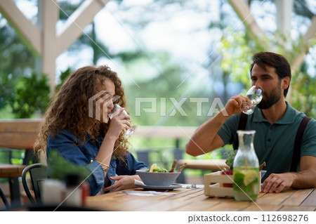 Young couple at date in restaurant, sitting on restaurant terrace, drinking water. Boyfriend and girlfriend enjoying springtime, having lunch outdoors. 112698276