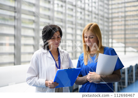 Two female doctors discussing test results, patients diagnosis. walking across corridor. Women in medicine science 112698467