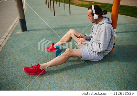 Athletic man in headphones, checking mobile app on his smartphone while resting after workout the urban sports ground 112698529