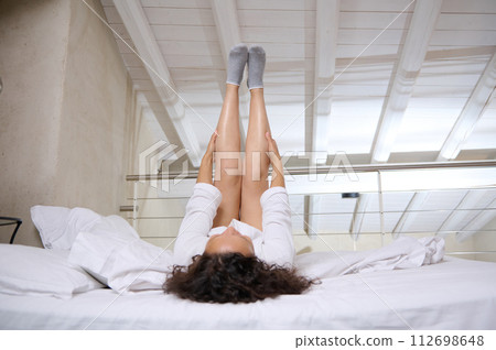 Young pretty woman stretching while wakes up in the morning, lying on bed on white bedsheets relaxed with legs raised up Young pretty woman stretching while wakes up in the morning, lying on bed on white bedsheets relaxed with legs raised up 112698648