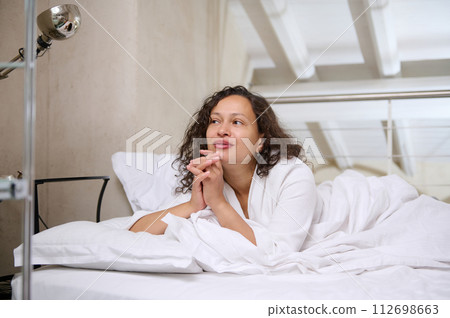 Happy positive woman smiling dreamily looking away, lying on the bed, enjoying her weekend morning while waking up 112698663