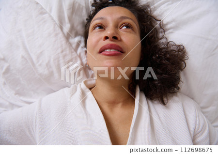 View from above smiling young adult woman waking up in the morning , lying on the bed at home bedchamber. 112698675