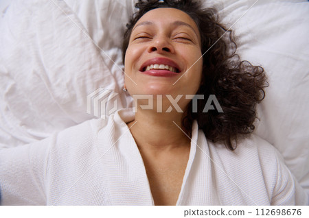 Close-up portrait of a smiling curly young woman smiling while waking up in the morning , lying on the bed 112698676